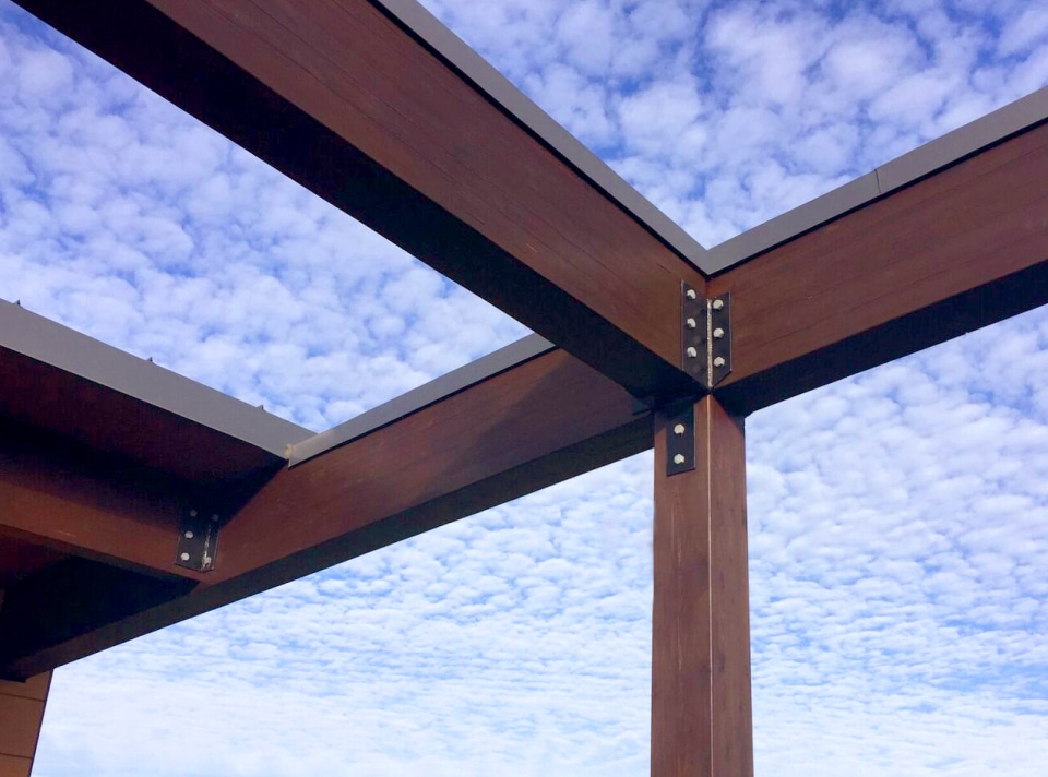 Des poutres en bois forment une structure ouverte sur un ciel rempli de nuages duveteux et épars à la station de pompage de Masson-Mascouche. Les poutres sont reliées par des supports métalliques, contrastant le bois brun riche avec le ciel bleu et blanc éclatant au-dessus de cette installation vitale d'eau potable.
