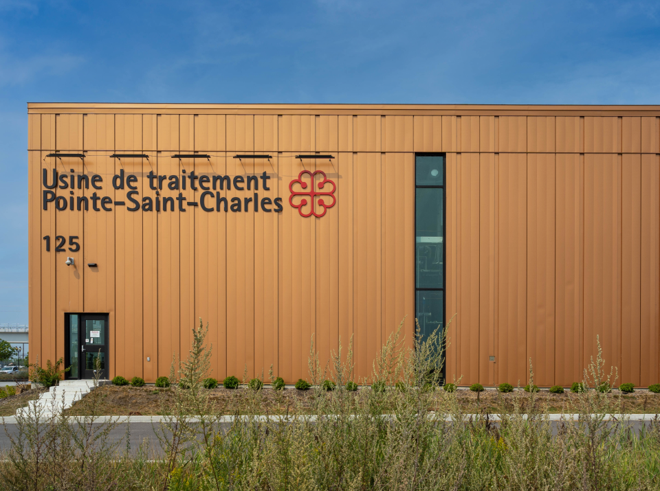 Un grand bâtiment industriel au revêtement métallique couleur rouille arbore un logo floral rouge à côté du texte « Usine de traitement Pointe-St-Charles » et du numéro « 125 ». De hautes herbes ondulent au premier plan sous un ciel bleu clair dans cette usine de traitement.
