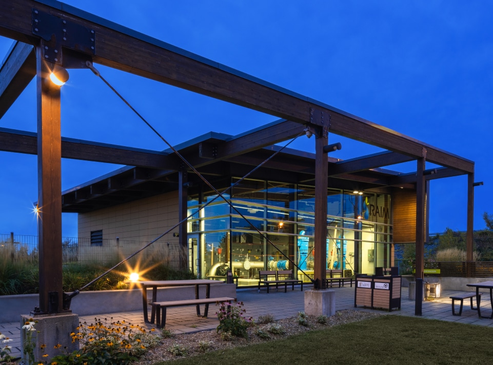 Bâtiment moderne avec de grandes baies vitrées illuminées au crépuscule. La structure, ressemblant à une station de pompage sophistiquée, présente des poutres en bois et un espace paysager avec des bancs et des plantes au premier plan. Le ciel bleu profond renforce l'atmosphère sereine du soir.