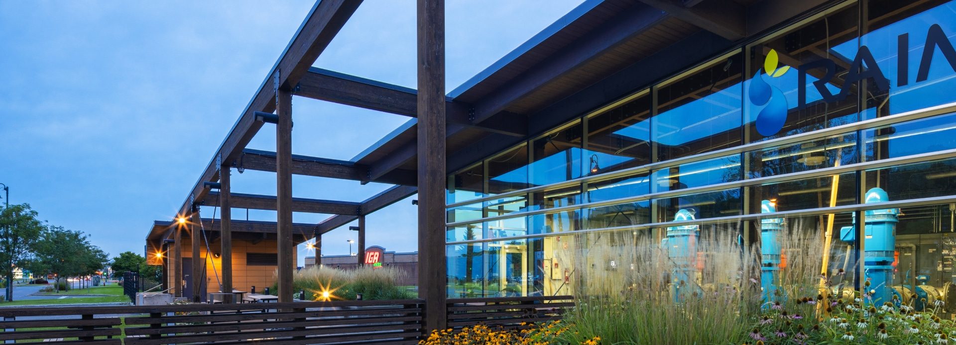 L'extérieur de l'édifice moderne, niché dans le quartier Masson-Mascouche, met en valeur de grandes fenêtres vitrées reflétant un ciel nuageux et des poutres en bois. Au premier plan, des plantes et des fleurs décoratives prospèrent près d'une station de pompage. Une vue partielle d'un logo orne la structure.