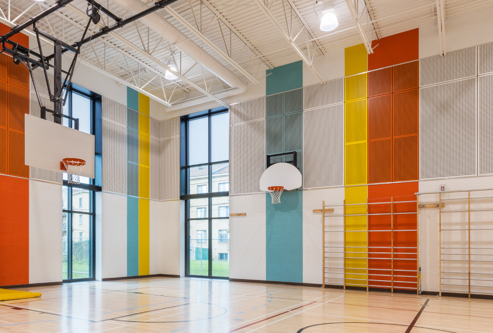 Le terrain de basketball intérieur de l'école Marc-Aurèle Fortin de Montréal arbore des murs colorés aux rayures verticales orange, bleu sarcelle, jaune et grise. Deux paniers sont installés sous la lumière naturelle provenant de hautes fenêtres, tandis que des équipements sportifs agrémentent le récent agrandissement du gymnase.