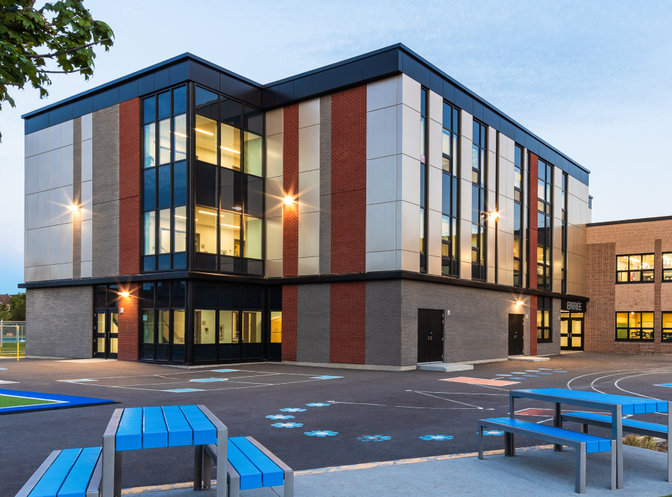 L'école Marc-Aurèle Fortin de Montréal se distingue par son design moderne sur trois étages, ses grandes fenêtres vitrées et son mélange de briques et de panneaux gris. La cour de récréation peinte et les bancs en métal bleu-gris rehaussent son charme, tandis que les lumières du bâtiment projettent une lueur subtile sous le ciel clair, reflétant l'agrandissement récent.