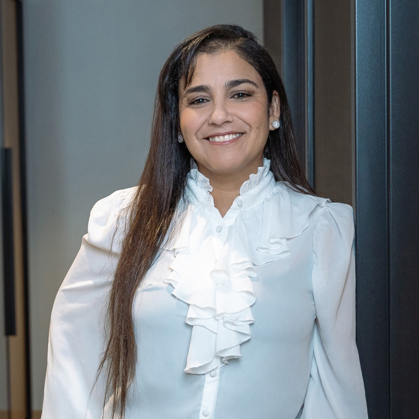 Sana Smida - Une femme aux longs cheveux noirs sourit et porte un chemisier blanc à volants sur le devant, comme si elle était sur le point d'occuper le devant de la scène. Elle se tient dans un décor intérieur aux murs aux tons neutres, prête pour son moment sous les projecteurs, un véritable test de confiance et de style.