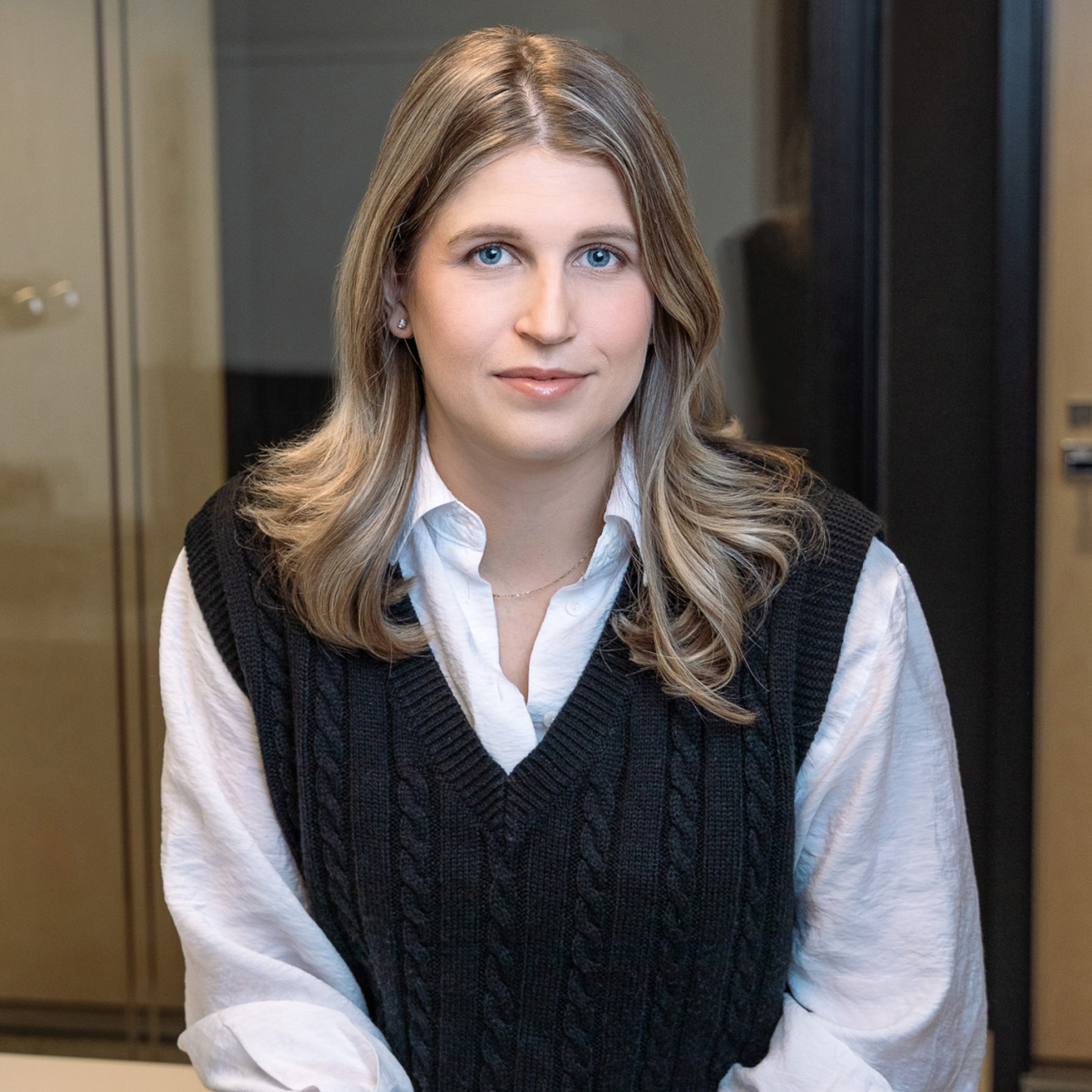Arianne Vigneault - Une personne aux cheveux longs et blonds porte une chemise blanche sous un gilet noir. Elle est assise dans un bureau aux murs en verre. La personne regarde directement la caméra avec une expression neutre.