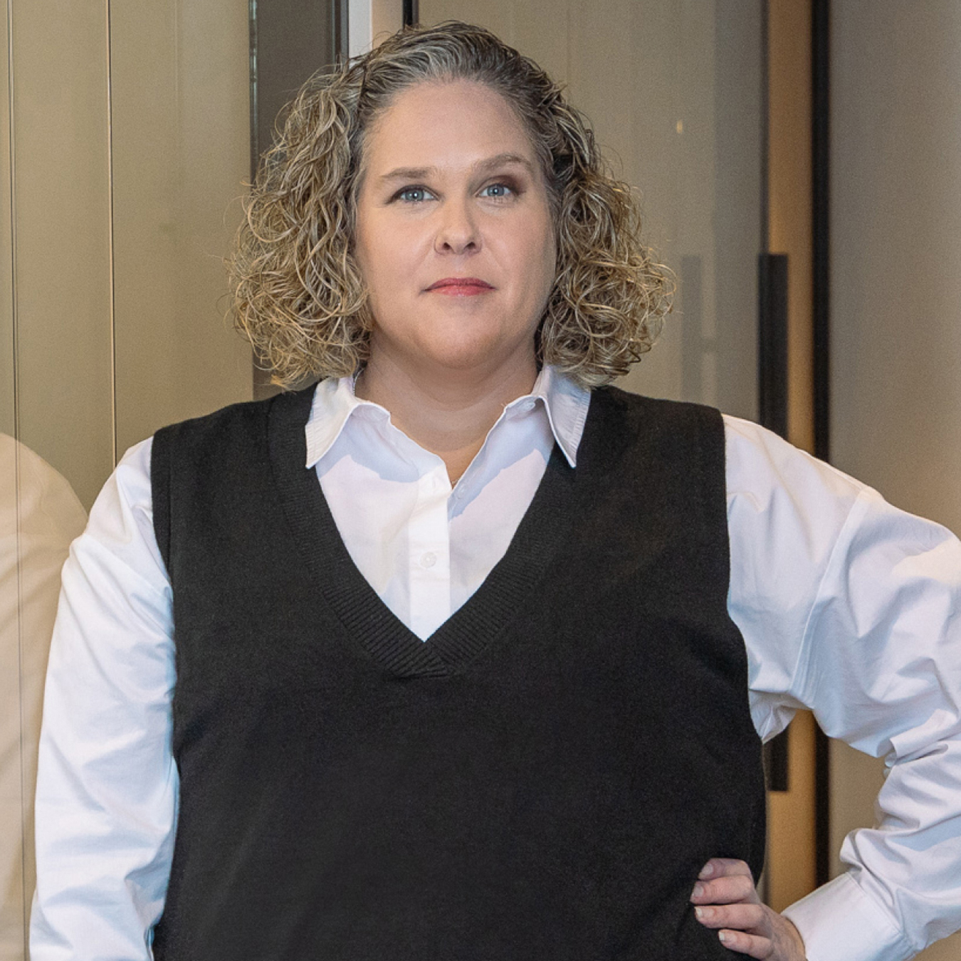 Catherine Larente - Une personne aux cheveux ondulés jusqu'aux épaules se tient debout, confiante, à l'intérieur. Elle porte une chemise blanche et un gilet noir. L'arrière-plan présente des panneaux de verre et une porte.