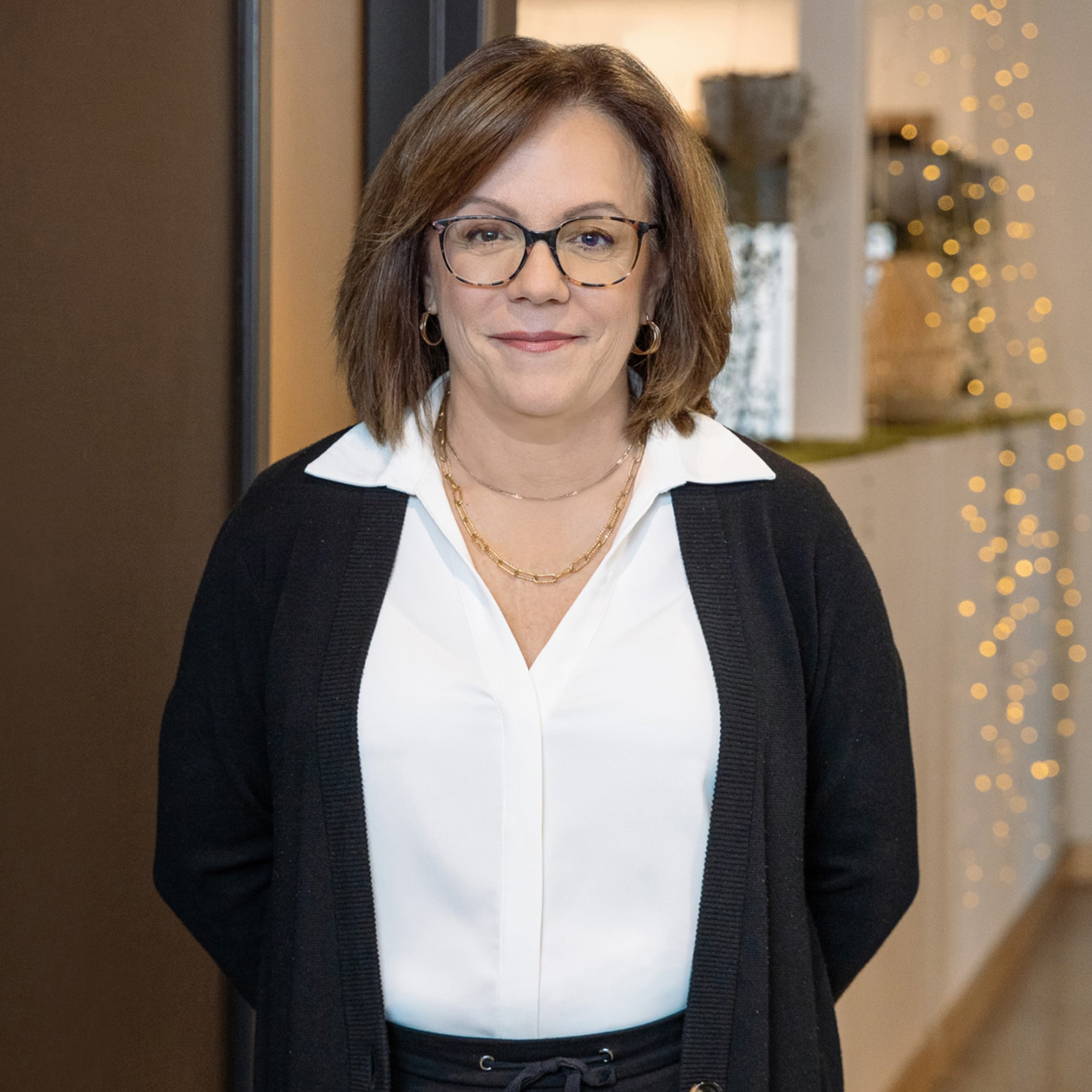 DIANE LANGELIER - Une personne aux cheveux châtains mi-longs et portant des lunettes porte un chemisier blanc et un cardigan noir. Elle se tient debout dans une pièce avec un doux effet bokeh provenant de guirlandes lumineuses en arrière-plan.