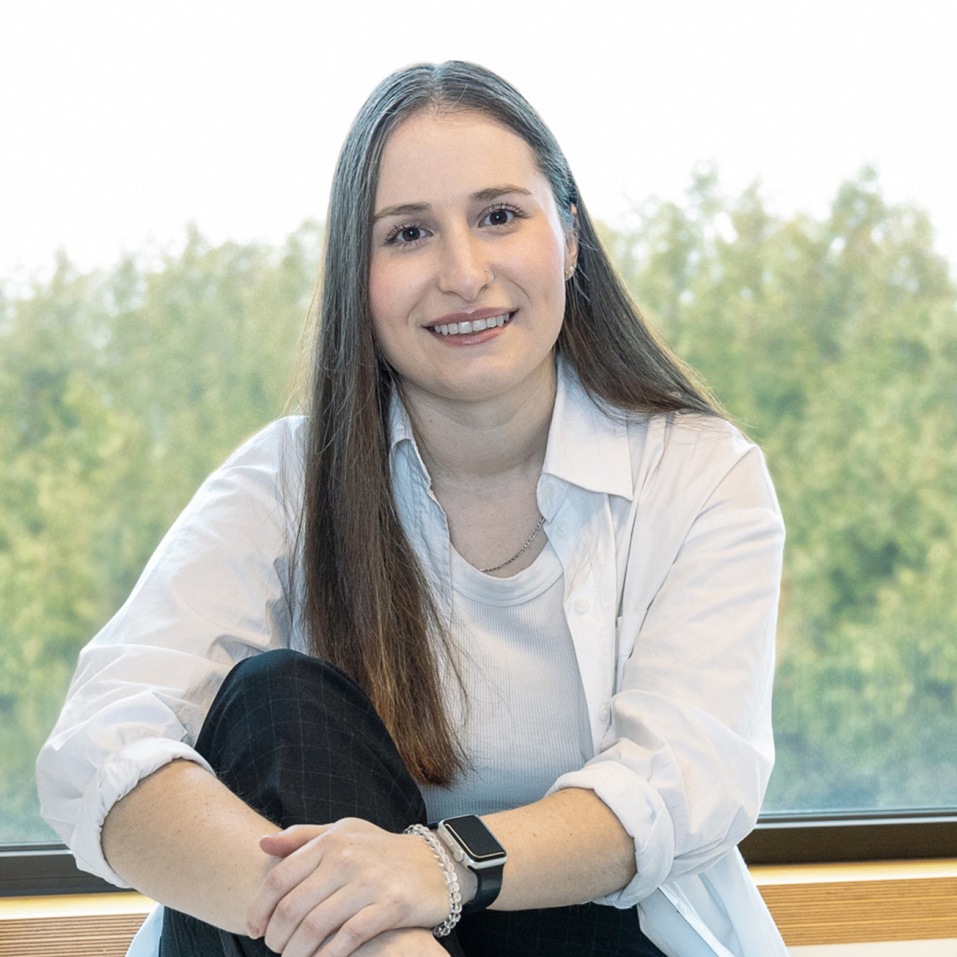 Jade Côté - Une personne aux cheveux longs est assise et sourit, vêtue d'une chemise blanche sur un haut blanc et d'un pantalon noir, avec une montre connectée et un bracelet. Elle est assise près d'une fenêtre, avec vue sur des arbres verts en arrière-plan.