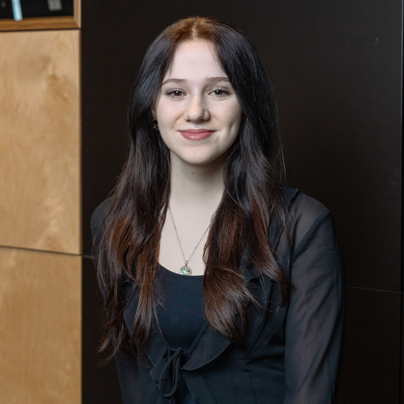 Juliette Gadoury - Personne aux cheveux longs et noirs, portant un haut noir et un collier pendentif, souriant doucement. Ils se tiennent devant un fond en bois et noir.