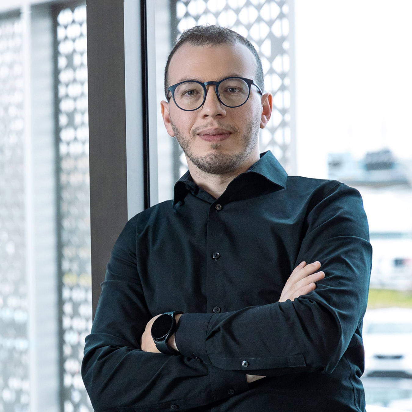 Omar Abada - Une personne portant des lunettes et une chemise noire se tient debout, les bras croisés, souriant légèrement, devant une fenêtre avec un écran à motifs. L'arrière-plan est flou.