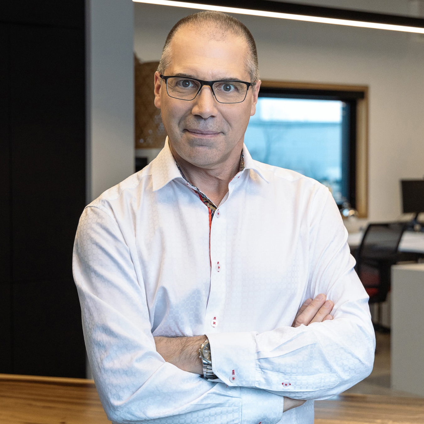 Pascal Roffi - Un homme aux cheveux courts et aux lunettes porte une chemise blanche boutonnée. Il se tient debout, les bras croisés, dans un espace de bureau moderne, avec un bureau, une chaise et une grande fenêtre en arrière-plan.