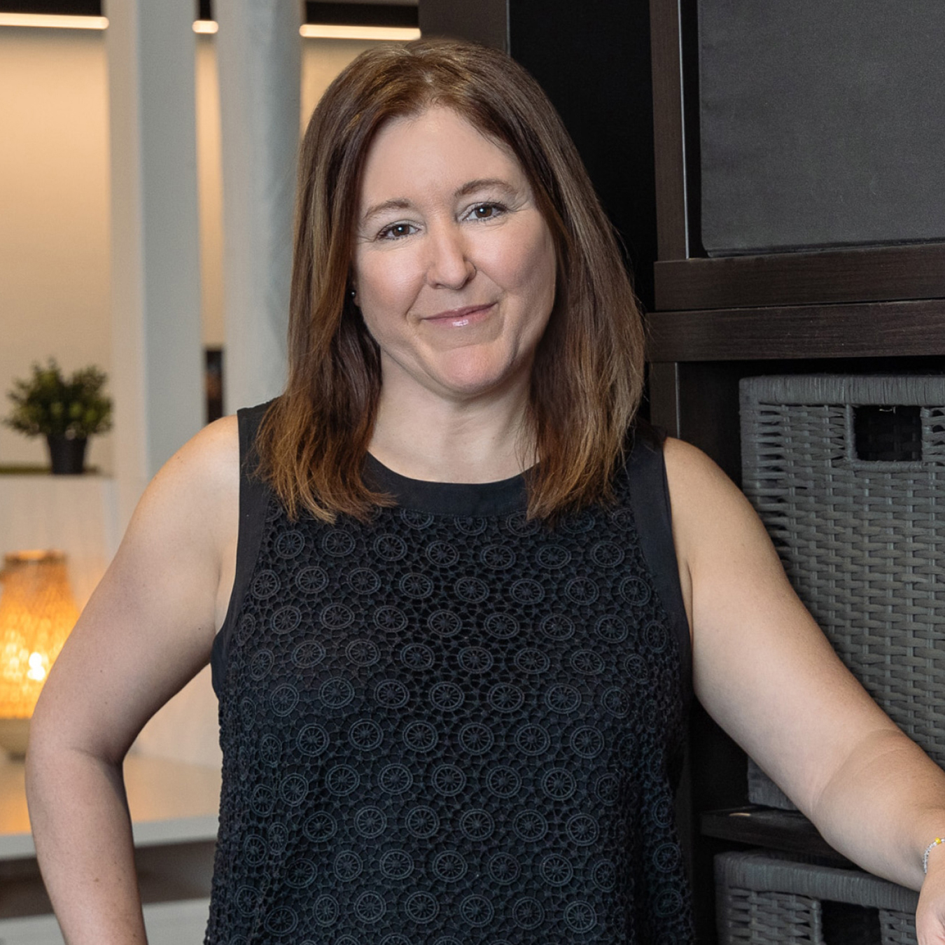 Valérie Laforme - Une femme aux cheveux châtains mi-longs, vêtue d'un haut noir sans manches, s'appuie nonchalamment contre un meuble. Elle sourit doucement. L'arrière-plan montre un intérieur moderne avec un éclairage tamisé et des éléments décoratifs.