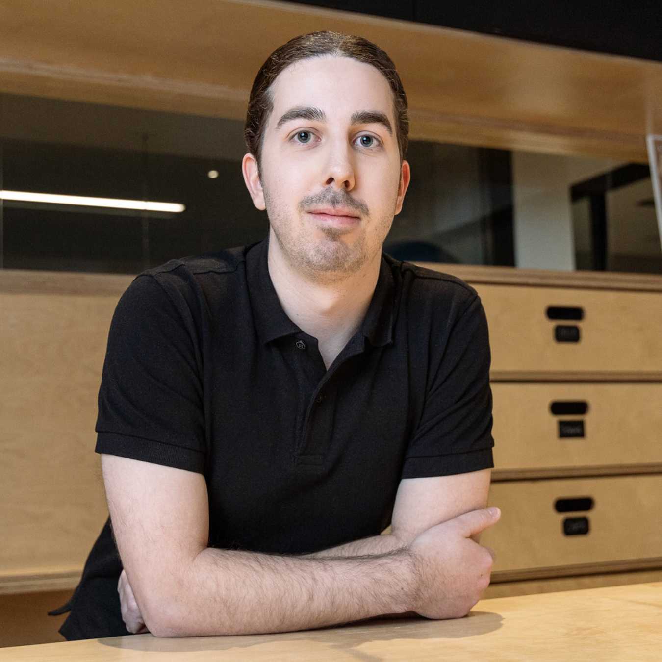 William Gagné - Une personne aux cheveux noirs courts, vêtue d'un polo noir, est assise à une table en bois, les bras croisés. L'arrière-plan montre des tiroirs en bois et une partie d'un espace de bureau moderne.