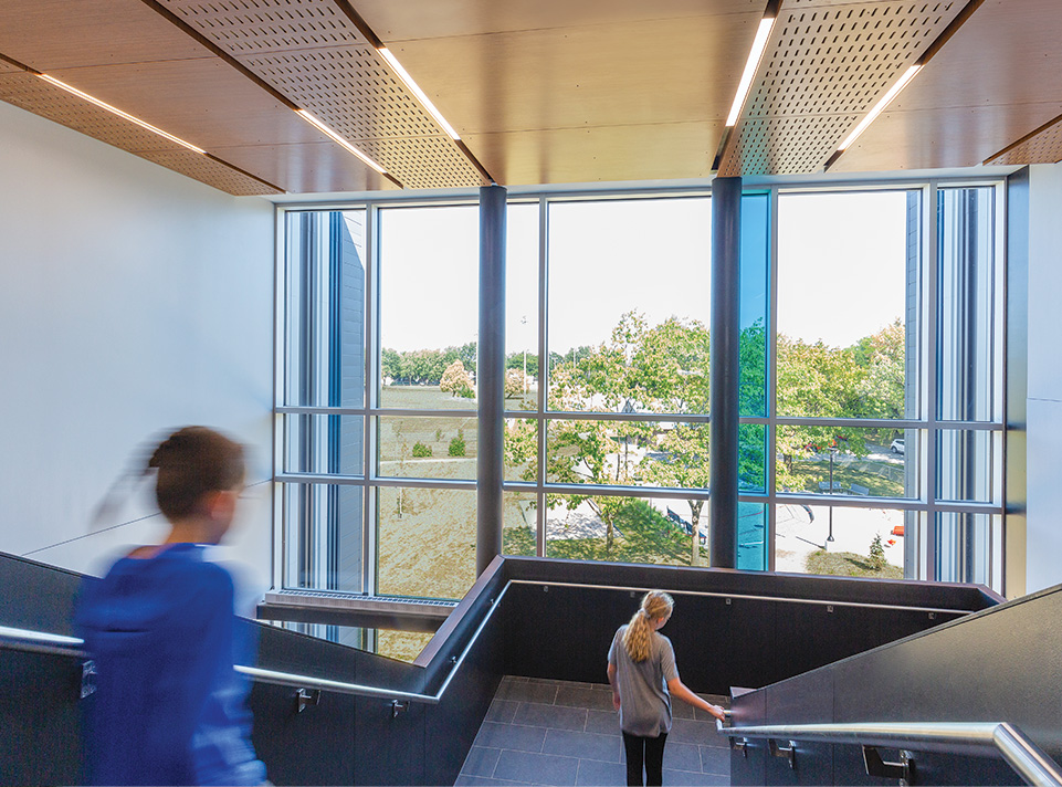 Nouvelle école des Aventuriers. Deux enfants descendent les escaliers devant une large fenestration laissant entrevoir des arbres. La fenestration est ponctuée d'un vitrail bleu et laisse passer la lumière du jour. le plafond texturé est incrusté de néons.
