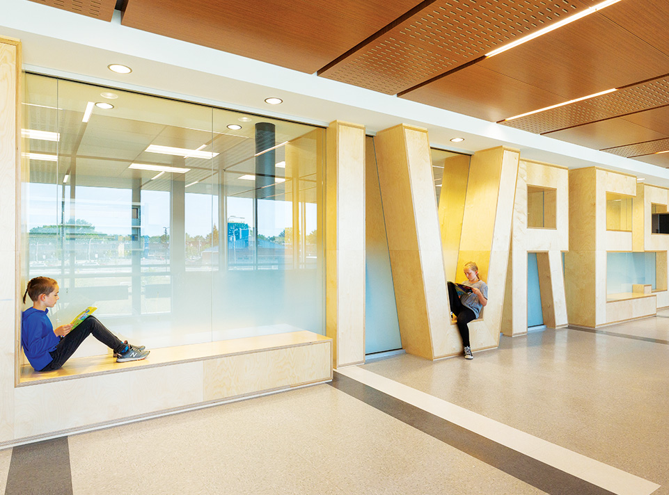 Nouvelle école les Aventuriers. Espace cloisonnant la bibliothèque donnant sur le corridor. Les lettres L-I-V-R-E-S en panneaux de bois blond sont judicieusement formées en 3 dimensions et éclairées par des lumières encastrées pour structurer cette cloison. Des enfants sont assis dans la structure du mot ''livres'' pour faire de la lecture et pour démontrer l'utilité fonctionnelle de la cloison symbolique. Le reflet de la fenestration donne des effets bleutés à l'ensemble. Le plafond texturé est éclairé par des néons.