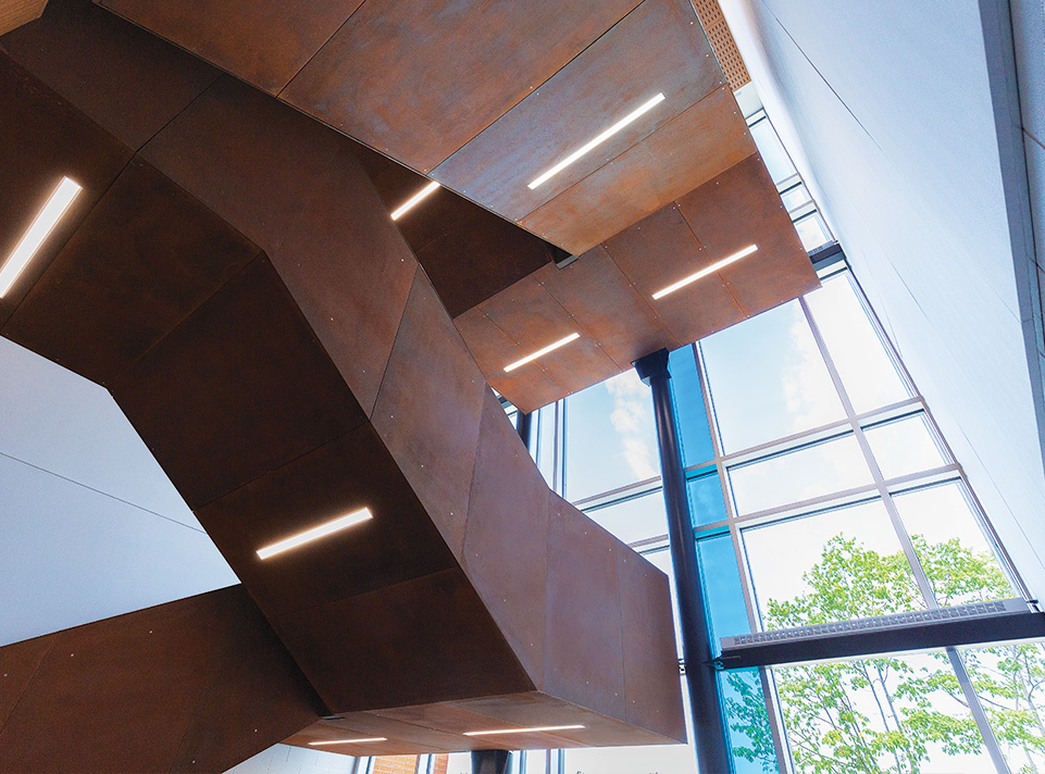Nouvelle école des Aventuriers. Détails du dessous de la cage d'escalier en métal oxydé faisant contraste avec la fenestration incluant un vitrail bleu. Les angles dynamisent la prise de vue. L'éclairage extérieur et des néons intégrés dans la structure de l'escalier illuminent l'espace.