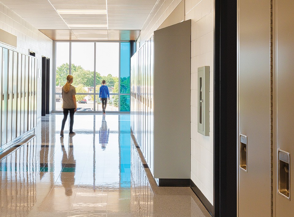 Nouvelle école des Aventuriers vue d'un corridor du second étage, très lumineux. La fenestration laisse entrer la lumière du jour avec une insertion d'un vitrail bleu se reflétant au sol. Deux enfants déambulent dans le couloir ou des vestiaires sont visibles de chaque cotés du corridor.