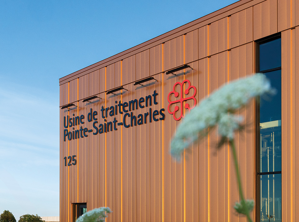Usine de pré-traitement des eaux souterraines Pointe-St-Charles à Montréal. Vue rapproché de la signalisation appliquée sur le bâtiment. Le logo de la Ville de Montréal est très apparent ainsi que l'adresse et la nomination de l'usine. En premier plant, une floraison floue donne un aspect poétique à la photographie. Lumière douce sur un ciel bleu de fin d'après-midi en été.