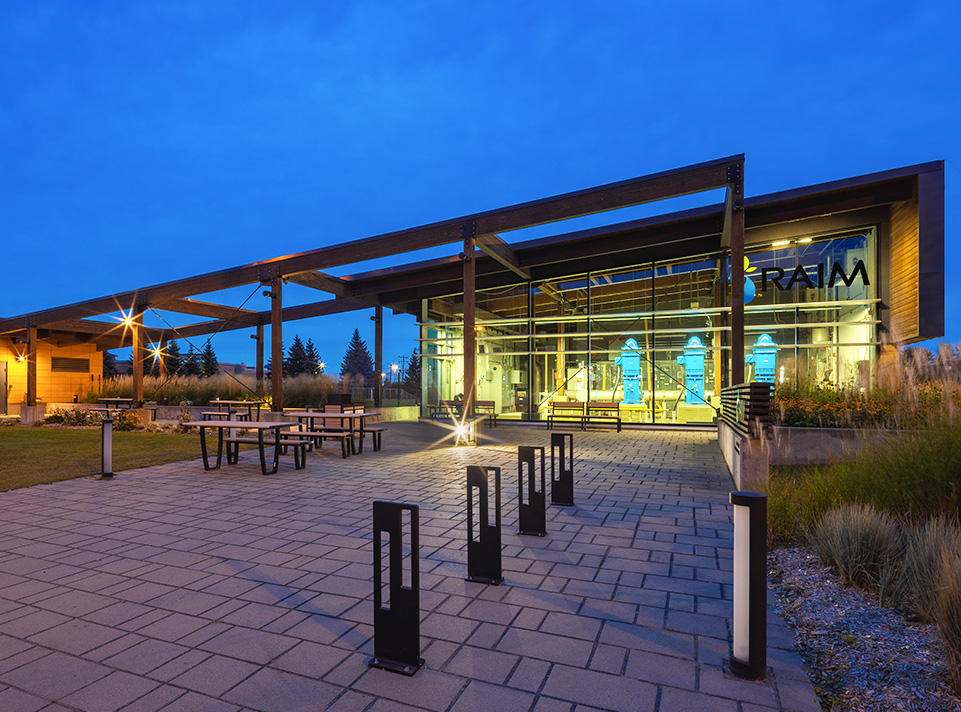 Station de pompage d'eau potable Masson-Mascouche. Aire de repos avec éclairage urbain, tables de pique-nique, végétation et pavé uni au sol. Une grande pergola relie les deux bâtiments de la station de pompage. La signalisation RAIM est très visible. Éclairage de fin du jour, en été.