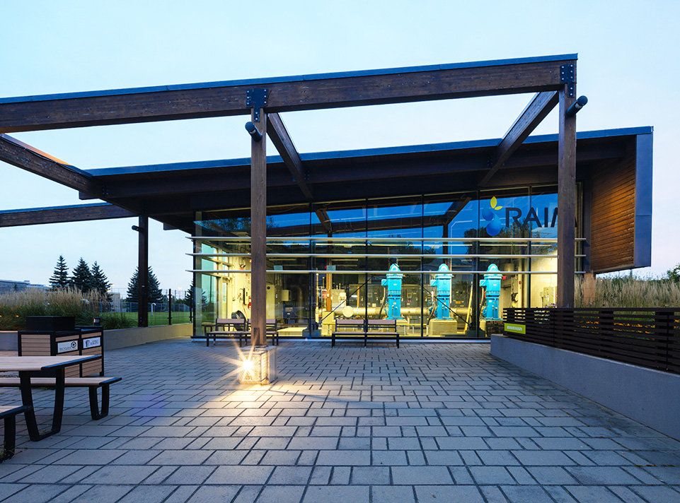 Station de pompage d'eau potable Masson-Mascouche. Aire de repos avec éclairage urbain, tables de pique-nique, végétation et pavé uni au sol. Une grande pergola est intégrée au bâtiment de la station de pompage. La signalisation RAIM est très visible. Éclairage de fin du jour, en été.