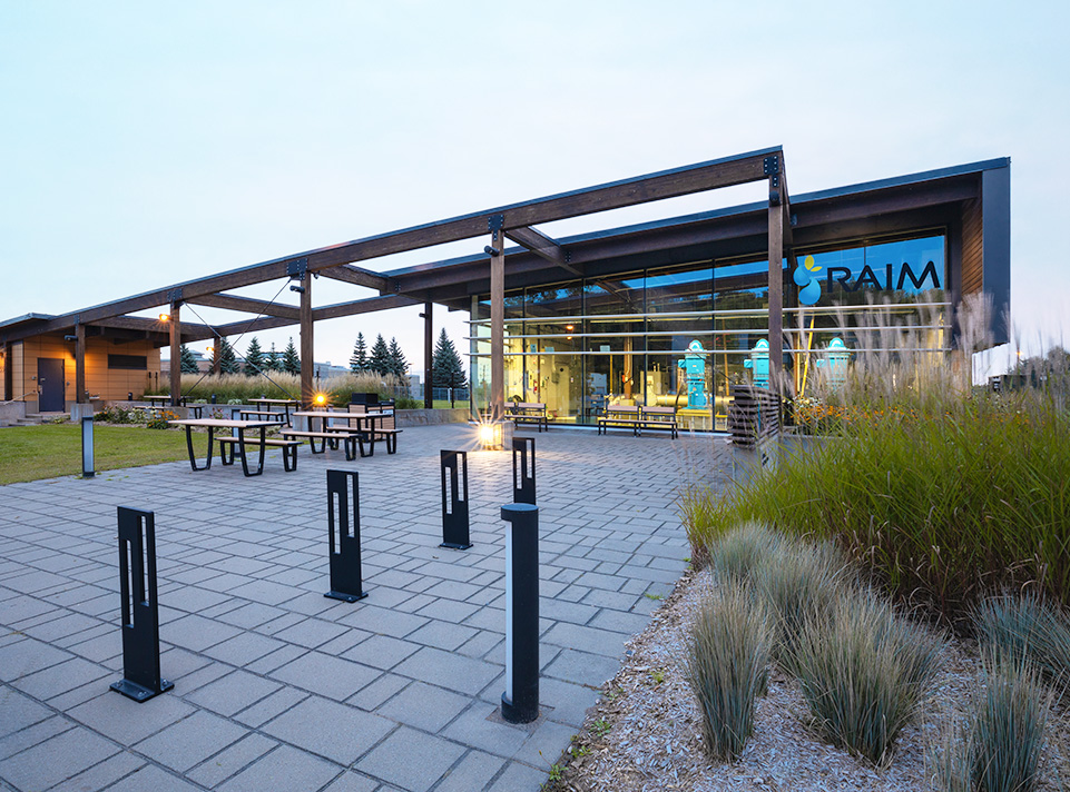 Station de pompage d'eau potable Masson-Mascouche. Aire de repos avec éclairage urbain, tables de pique-nique, végétation et pavé uni au sol. Une grande pergola relie les deux bâtiments de la station de pompage. La signalisation RAIM est très visible. Éclairage de fin du jour, en été.