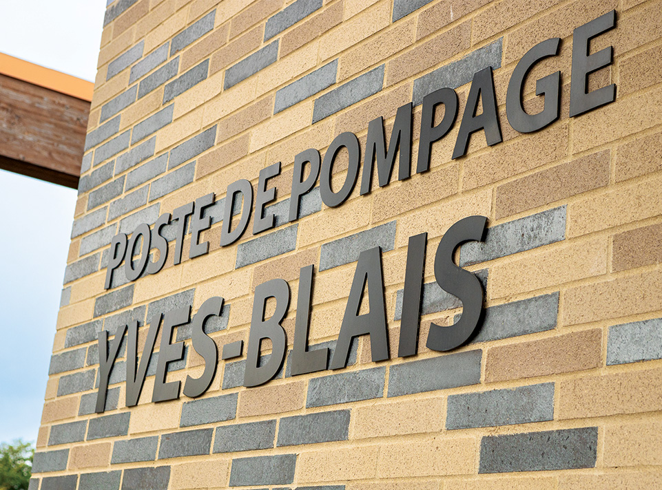 Station de pompage Yves-Blais - Pôle de la croisée. La station est située à Terrebonne, dans le secteur Lachenaie. Mur de brique avec variations de couleurs passant du noir, gris, brun et sable. La signalisation en lettres noires est appliquée en caractères majuscules '' POSTE DE POMPAGE YVES-BLAIS ''. On voit une poutre se détachant du ciel en plein jour.