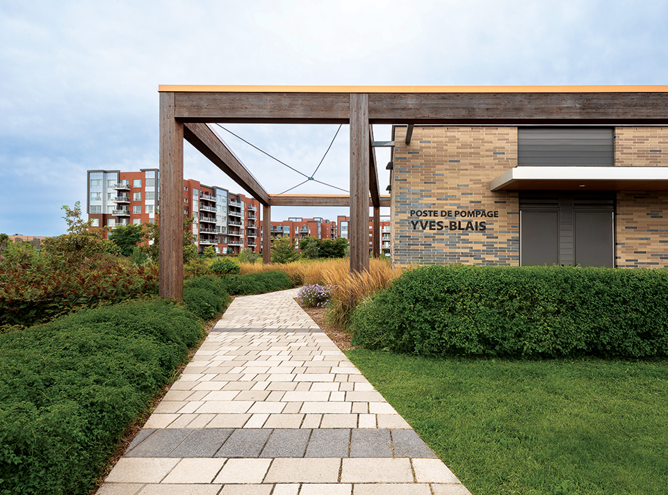 Station de pompage Yves-Blais - Pôle de la croisée. La station est située à Terrebonne, dans le secteur Lachenaie. Quartier d'habitation de type condominiums en brique rouille en arrière plan, se découpant sur un ciel gris bleu. On voit une allée de pavé qui passe sous la pergola intégrée à la station de pompage. L'aménagement paysager est en avant plan et rehausse la beauté du bâtiment aux tons de briques gris et sable. Éclairage de jour en été.