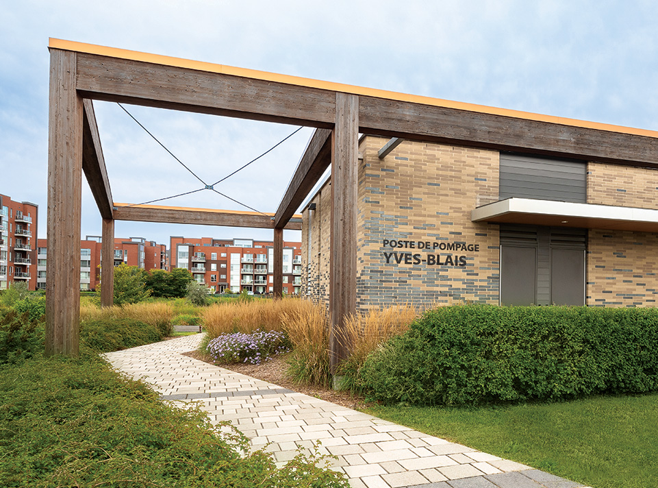 Station de pompage Yves-Blais - Pôle de la croisée. La station est située à Terrebonne, dans le secteur Lachenaie. Quartier d'habitation de type condominiums en brique rouille en arrière plan, se découpant sur un ciel gris bleu. On voit une allée de pavé qui passe sous la pergola intégrée à la station de pompage. L'aménagement paysager est en avant plan et rehausse la beauté du bâtiment aux tons de briques gris et sable. Éclairage de jour en été.