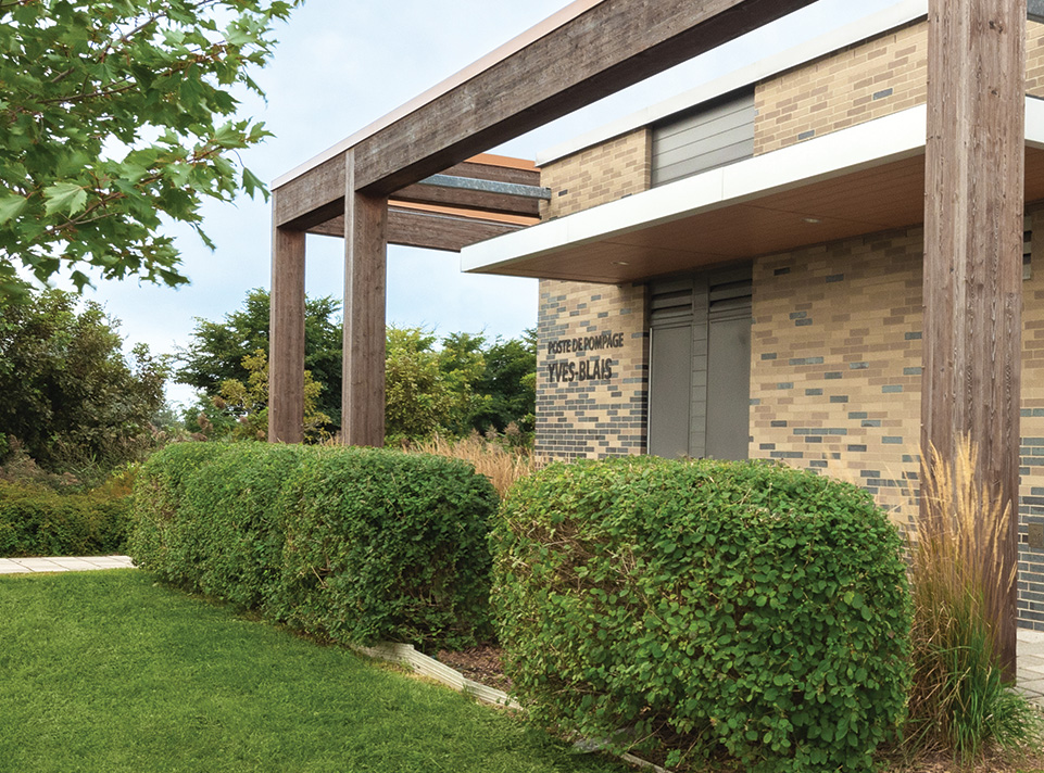 Station de pompage Yves-Blais - Pôle de la croisée. La station est située à Terrebonne, dans le secteur Lachenaie. Vue latérale de la façade de brique encadrée de la structure de bois faisant office de pergola, valorisant l'aménagement paysager luxuriant. On voir la signalisation identifiant la bâtisse POSTE DE POMPAGE YVES-BLAIS. Ciel bleu-gris éclairage de jour, de nombreux arbres verts à l'horizon. Période estivale.