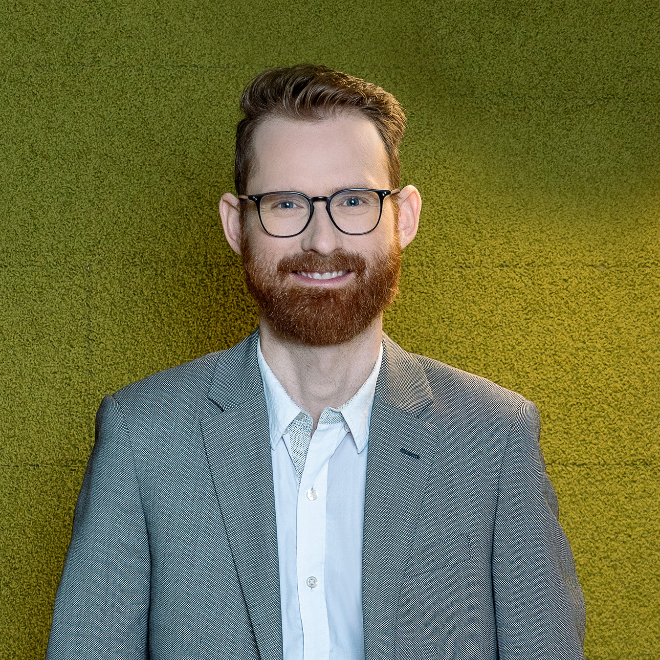 Frédéric Perron - Un homme aux cheveux brun roux et à la barbe, portant des lunettes et un costume gris avec une chemise blanche, se tient devant un mur vert texturé.