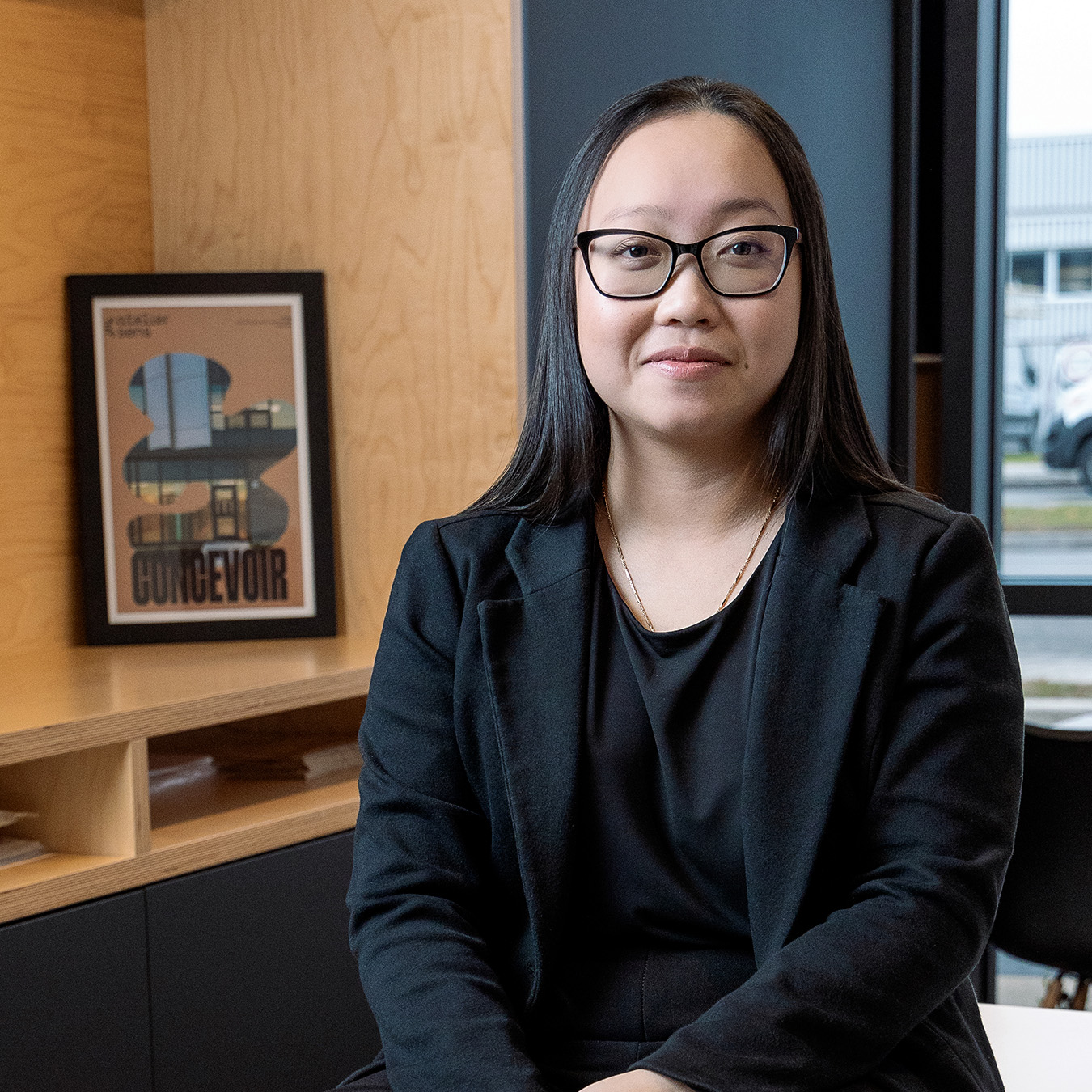 Jennifer Lai - Une personne aux cheveux longs et noirs et portant des lunettes sourit légèrement, vêtue d'un blazer noir sur un haut noir. Elle se tient dans une pièce avec des étagères en bois et une armoire sombre en arrière-plan.