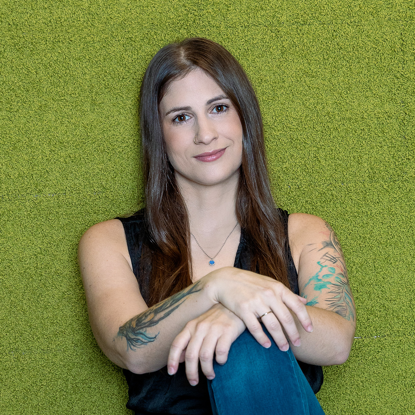 Rachel Mantha - Une femme aux longs cheveux bruns et aux tatouages sur les deux bras est assise sur un fond vert texturé. Elle porte un haut noir sans manches et un jean, souriant doucement avec ses mains jointes sur ses genoux.