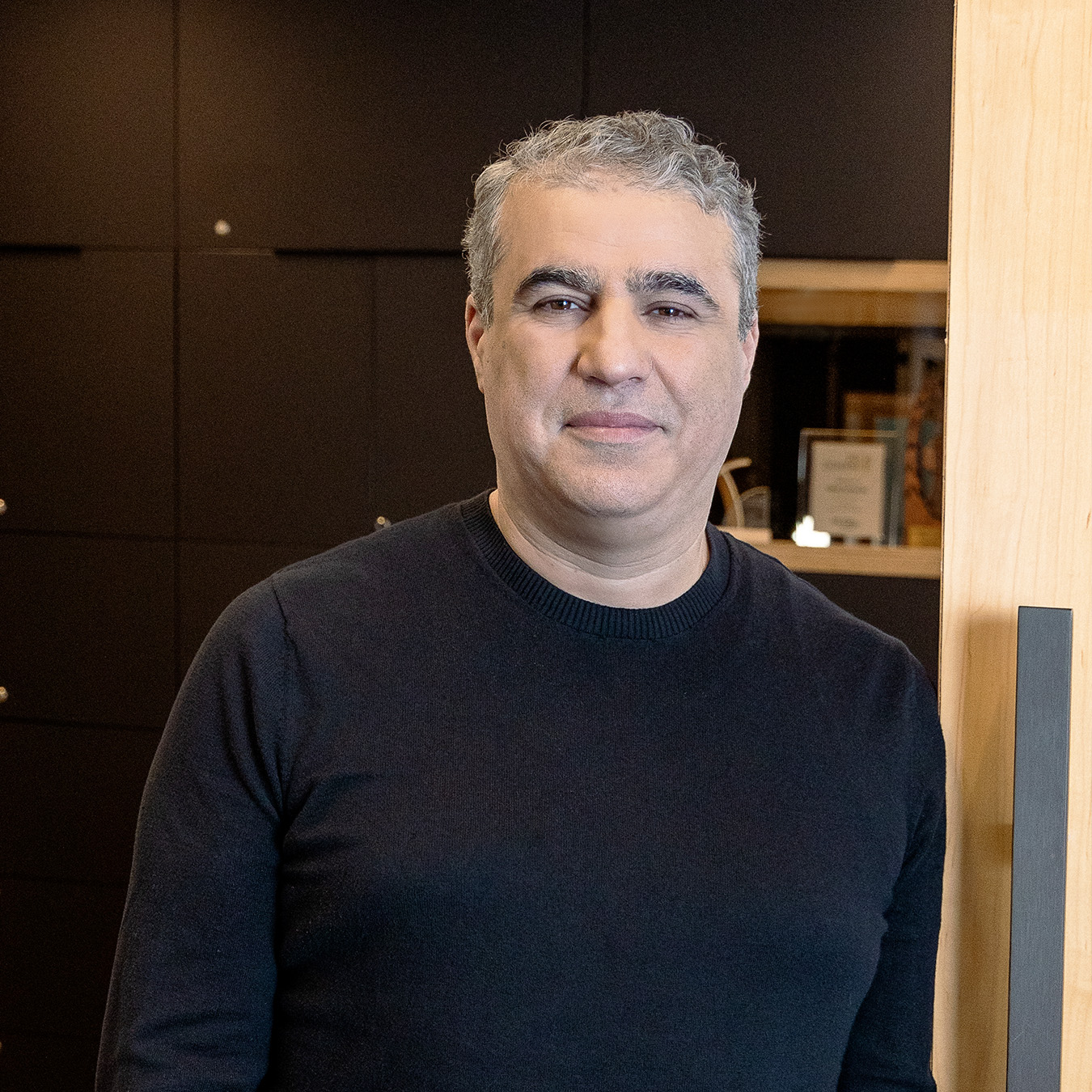 Salim Mohdeb. Une personne aux cheveux gris, portant un pull noir, se tient à l'intérieur près d'un mur lambrissé. L'arrière-plan présente des murs sombres ornés d'objets encadrés.
