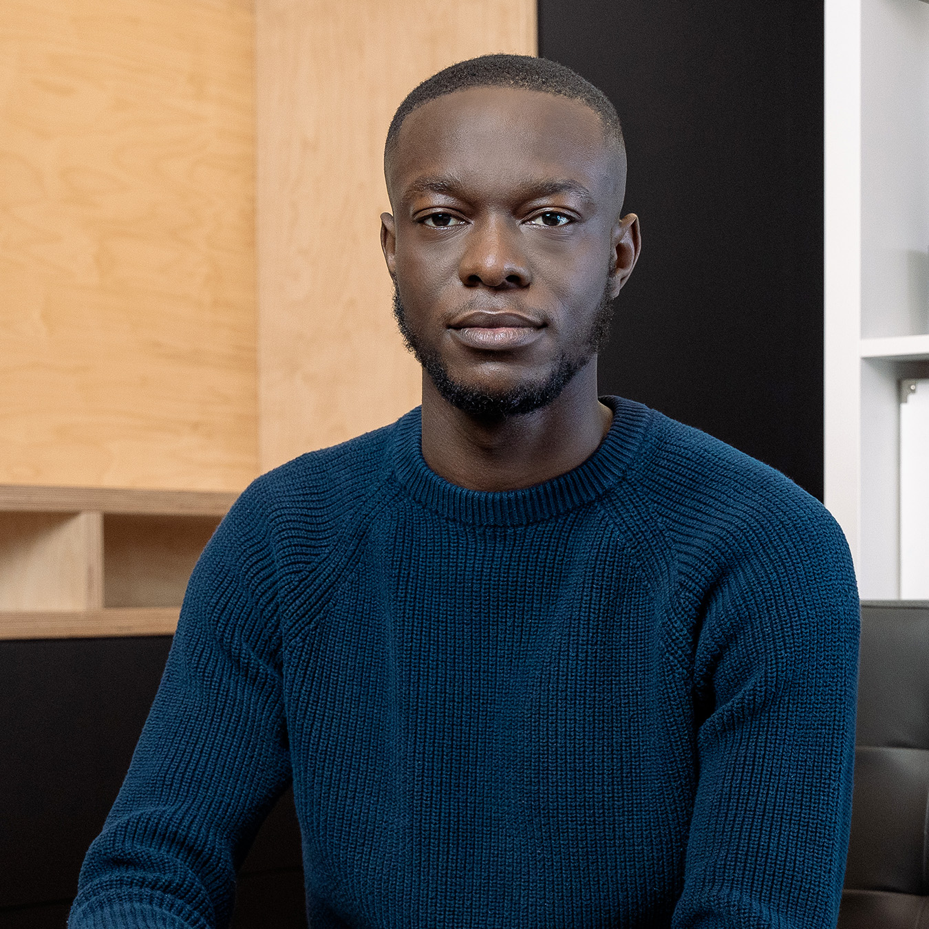 Astrid Fopa - Un jeune homme à la peau noire aux cheveux très courts et portant un pull bleu foncé se tient sur un fond corporatif.
