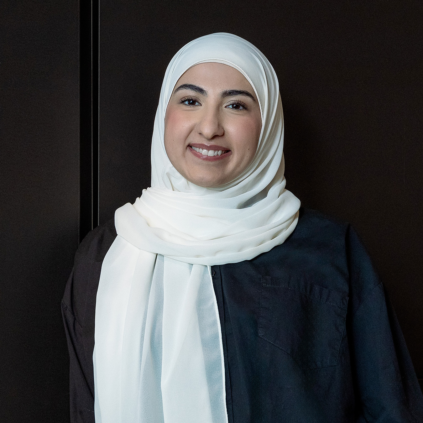 Sally Abd-Ali - Une jeune femme souriante aux cheveux dissimulés par un voile la drapant portant un chemisier noir se tient sur un fond noir.